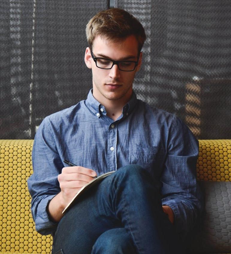 Joven con lentes sentado en un sillon escribiendo en un cuaderno.